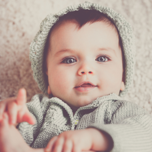 Baby playing with feet while looking forward wearing a hat.